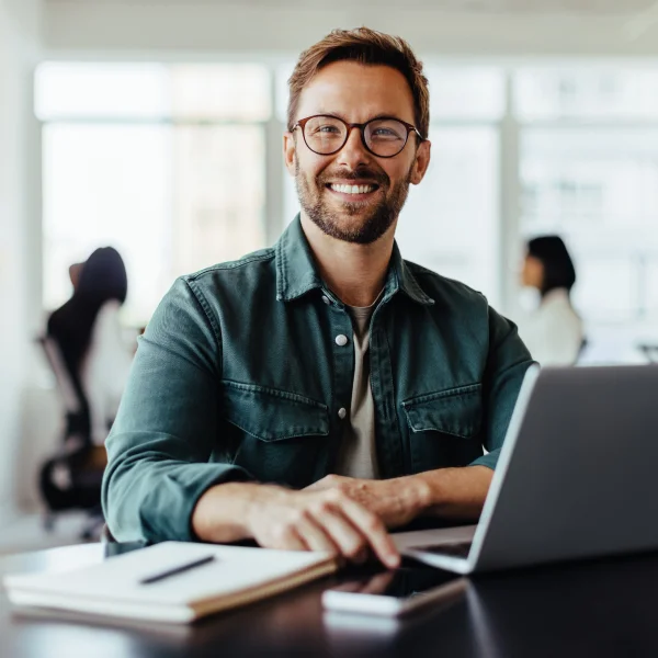 Un homme souriant portant des lunettes est assis à un bureau avec un ordinateur portable devant lui.